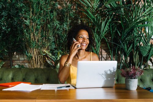 smiling-young-woman-talking-mobile-phone-with-laptop-documents-pen-wooden-table